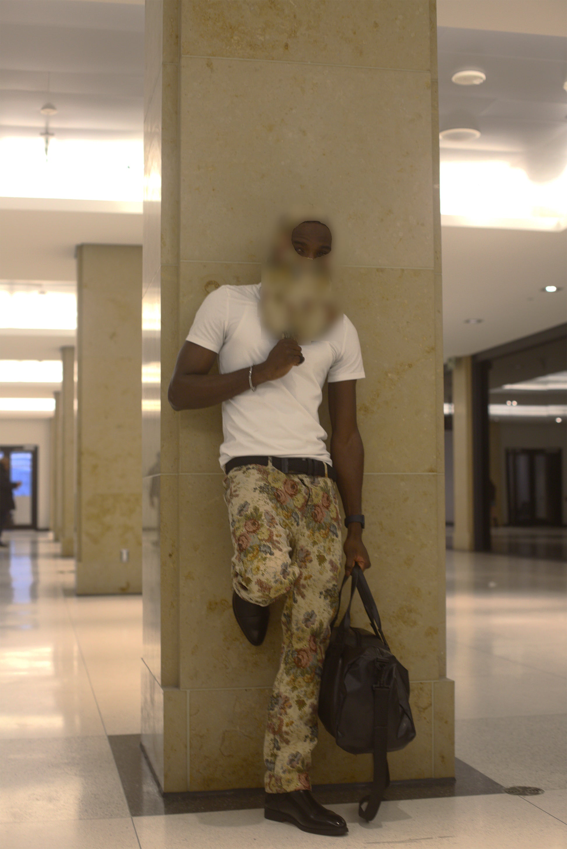 A man posing in a modern indoor lobby, wearing a white t-shirt, beige floral print pants, a matching face covering, and holding a black duffel bag.